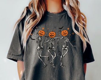 May include: A dark gray t-shirt with a graphic of three skeletons dancing with pumpkin heads. The skeletons are wearing black and white, and the pumpkins are orange with black stems. The background is a dark gray with white stars and bats.