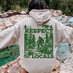 May include: A beige hoodie with a green graphic print of a Bigfoot in a forest setting. The graphic includes the text "RESPECT the LOCALS".