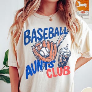 May include: Ivory-colored t-shirt with a baseball-themed graphic. The design features a baseball, glove, and a drink with ice, along with the text "BASEBALL AUNTS CLUB" in blue and red lettering.