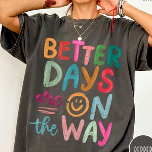 May include: A dark gray t-shirt with a colorful graphic that says "Better Days are on the Way" with a smiley face.