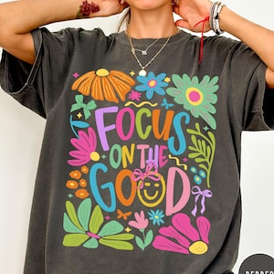 May include: Dark grey t-shirt featuring a vibrant floral design and the phrase "FOCUS ON THE GOOD". The design incorporates flowers in various colours, a smiling face, and bows. The t-shirt has a relaxed fit.