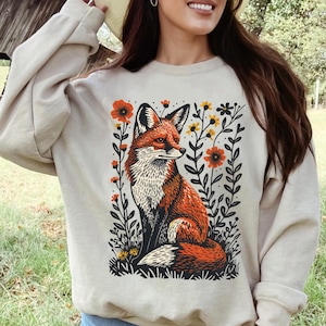 May include: A beige sweatshirt with a graphic print of a red fox sitting in a field of flowers. The flowers are in shades of red, yellow, and green. The text "SAND" is printed on the bottom left corner of the image.