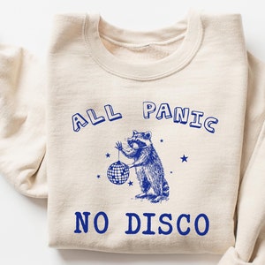 May include: A beige sweatshirt with a blue graphic of a raccoon holding a disco ball. The text "ALL PANIC NO DISCO" is printed in blue on the sweatshirt.