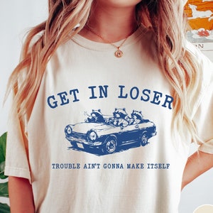 以下が含まれることがあります： クリーム色のTシャツに、青いグラフィックで描かれた、車に乗っているアライグマ3匹がプリントされています。「Get in Loser」というテキストがグラフィックの上に、「Trouble ain't gonna make itself」というテキストがグラフィックの下にあります。Tシャツには「アイボリー」というラベルが付いています。