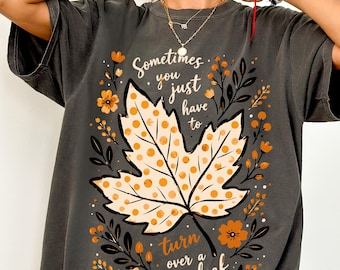 Puede incluir: Una camiseta gris carbón con un gráfico central de una hoja de arce con lunares naranjas. El diseño incluye detalles florales en naranja y negro, junto con el texto: Sometimes you just have to turn over a new leaf. Camiseta de manga corta.