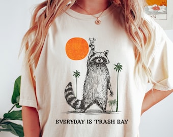 May include: A cream colored t-shirt with a graphic of a raccoon holding a drink in one hand and standing in front of a palm tree and a setting sun. The text "EVERYDAY IS TRASH DAY" is printed below the graphic. The t-shirt is available in the color ivory.