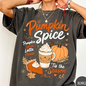 Puede incluir: Una camiseta gris carbón con un estampado gráfico que incluye el texto Pumpkin Spice y Tis the Season. El diseño muestra un latte de calabaza, una porción de tarta de calabaza y una calabaza entera, decorado con estrellas y hojas de otoño.