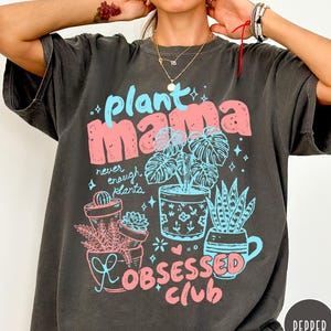 Puede incluir: Camiseta gris oscuro con las palabras "plant mama" en rosa y azul, junto con ilustraciones de plantas en macetas. Tambi&eacute;n aparece el texto "never enough plants" y "obsessed club".