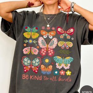 Puede incluir: Camiseta gris oscuro con un diseño de mariposas coloridas y el texto "Be Kind to all kinds". Las mariposas son de varios colores, incluyendo rojo, amarillo, rosa y azul. La camiseta tiene un corte holgado.