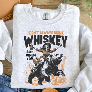 May include: A gray sweatshirt with a graphic print of a woman riding a bear. The text on the shirt reads "I don't always drink whiskey, but when I do..."
