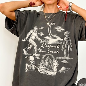Peut inclure: T-shirt gris foncé avec un motif graphique représentant Bigfoot, le monstre du Loch Ness, des extraterrestres et des ovnis. L'expression "Respect the Local" est imprimée en dessous. Le t-shirt a un aspect vintage.