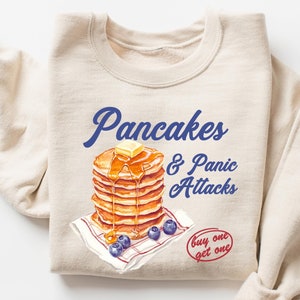 May include: A beige sweatshirt featuring a graphic of a stack of pancakes with butter and syrup, blueberries, and the text "Pancakes & Panic Attacks" in blue. The text "buy one get one" is in red.