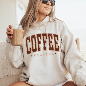 May include: A beige hoodie with a brown vintage-style graphic that says "COFFEE WEATHER" with stars on either side of the text. The model is holding a glass of iced coffee.