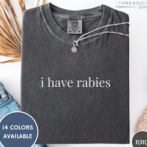 May include: Dark gray Comfort Colors t-shirt with the phrase "i have rabies" in white. A silver necklace with a circular pendant is on the shirt. The shirt is displayed with a blue circle that says "14 COLORS AVAILABLE".