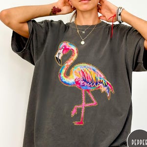 May include: Dark gray t-shirt featuring a colorful flamingo design. The flamingo is depicted in vibrant shades of pink, yellow, blue, and orange, with gold glitter accents. The shirt has a relaxed fit and short sleeves.