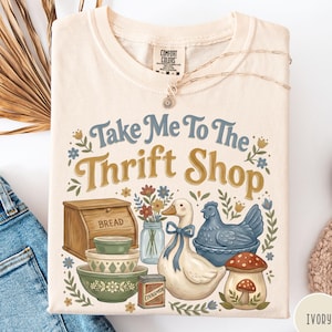 May include: Ivory-colored t-shirt with the text "Take Me To The Thrift Shop" in blue and gold. The graphic includes a bread box, bowls, a duck, a chicken, and a mushroom.