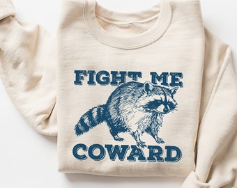 May include: A beige sweatshirt with a blue graphic of a raccoon and the text "FIGHT ME COWARD".
