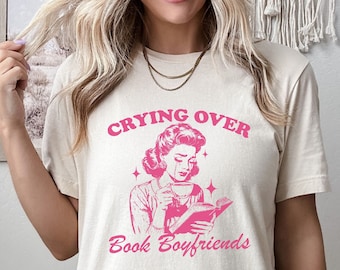 May include: A white t-shirt with the text "Crying Over Book Boyfriends" in pink lettering. A retro illustration of a woman with tears in her eyes holding a book and a cup of coffee is featured on the shirt.