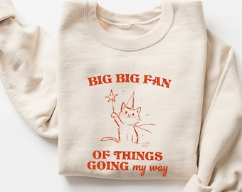 May include: A beige sweatshirt with a graphic of a cat wearing a party hat and holding a wand. The text "BIG BIG FAN OF THINGS GOING my way" is printed on the sweatshirt.