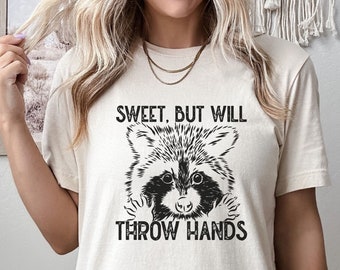 May include: A beige t-shirt with a black and white illustration of a raccoon with the text "Sweet. But Will Throw Hands".