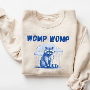 May include: A beige sweatshirt with a blue raccoon graphic and the text "WOMP WOMP" printed on the front.