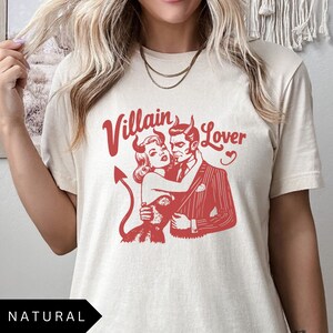 May include: A cream colored t-shirt with a red graphic of a devil couple. The text "Villain Lover" is above the couple.