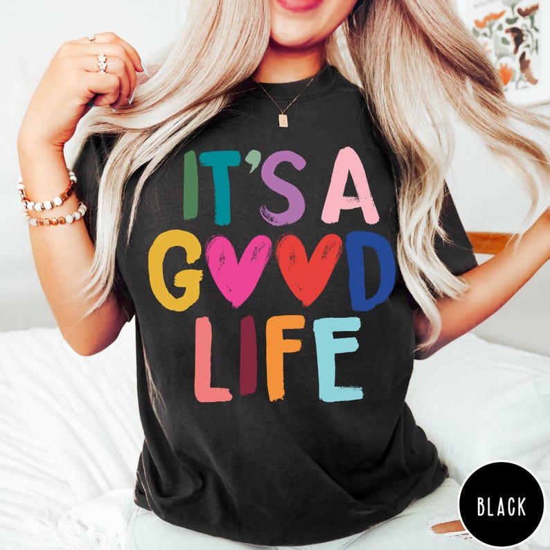 K&ouml;nnte beinhalten: Ein schwarzes T-Shirt mit kurzen &Auml;rmeln und dem mehrfarbigen Schriftzug "IT'S A GOOD LIFE" auf der Vorderseite. Die Buchstaben sind in verschiedenen leuchtenden Farben gehalten, darunter Gr&uuml;n, Blaugr&uuml;n, Lila, Pink, Gelb, Rot, Blau, Orange und Hellblau.
