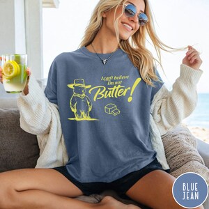 May include: A blue jean t-shirt with a yellow graphic of a bear wearing a cowboy hat and the text "I can't believe I'm not Butter!"