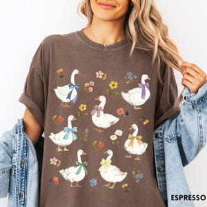 K&ouml;nnte beinhalten: Ein braunes T-Shirt mit einem Muster aus wei&szlig;en G&auml;nsen mit bunten Schleifen und floralen Akzenten. Die G&auml;nse sind in einem Rastermuster auf dem Shirt angeordnet.