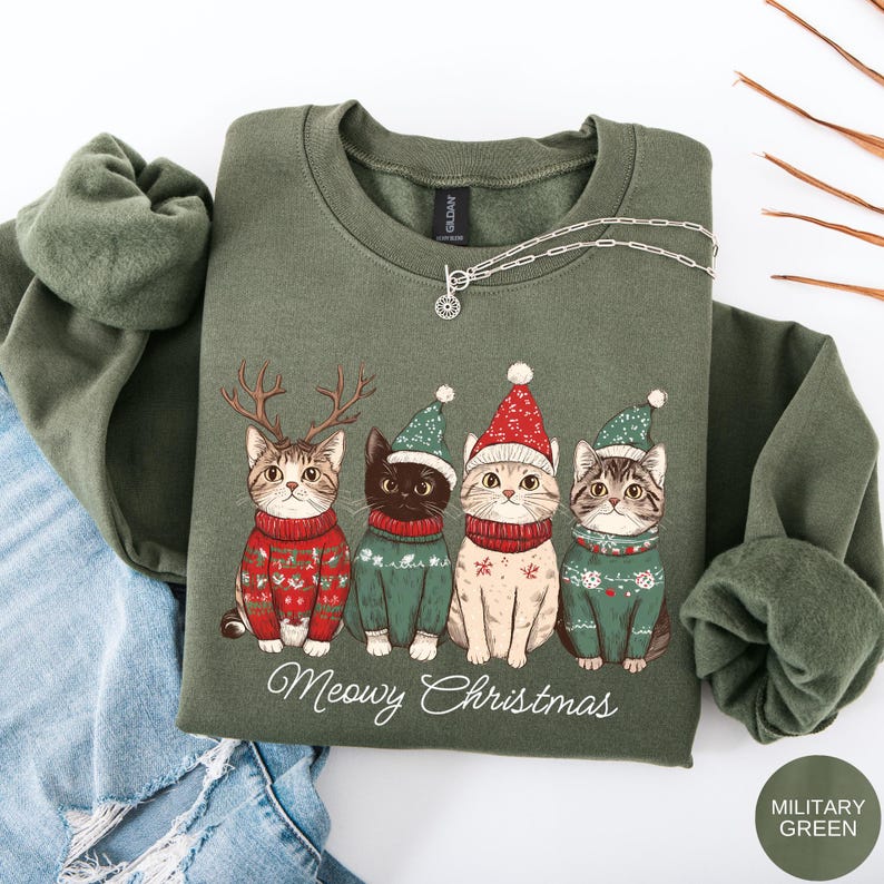May include: Olive green sweatshirt with a graphic of four cats wearing holiday sweaters and hats. The cats are tan, black, and grey, with the text "Meowy Christmas" below. A silver necklace is draped on the sweatshirt.