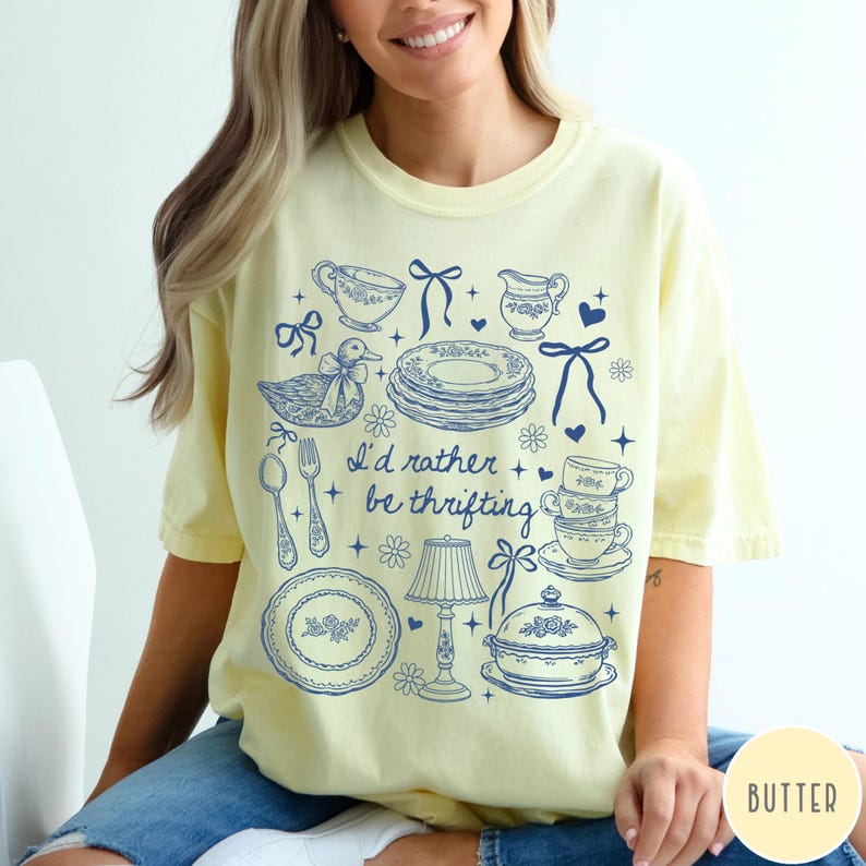 May include: Pale yellow t-shirt featuring a blue illustration of vintage crockery, a duck, and the text "I'd rather be thrifting." The design includes plates, teacups, a lamp, and cutlery. The shirt is a relaxed, casual style.