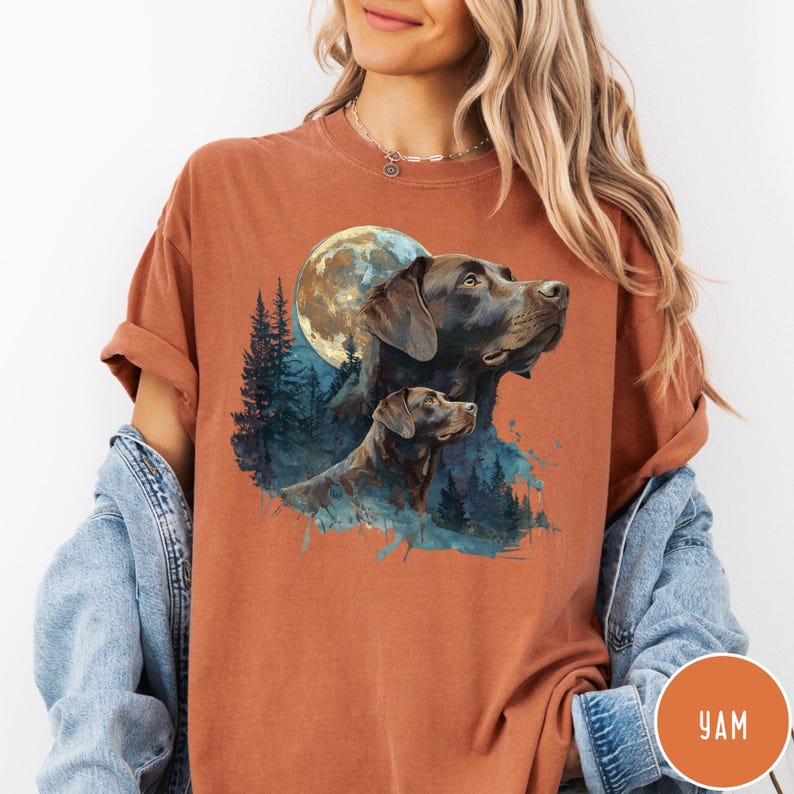 May include: Rust-colored t-shirt featuring a graphic of two Labrador Retrievers with a full moon and forest background. The dogs are depicted in shades of brown and black. The shirt has short sleeves and a relaxed fit.