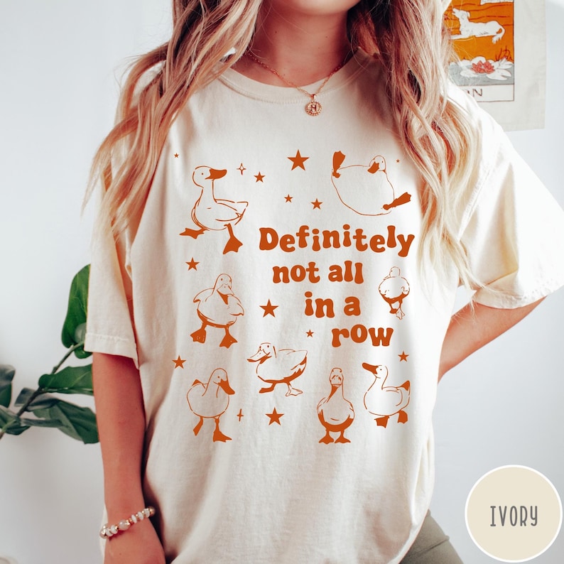 May include: Ivory t-shirt with a graphic of ducks in a row and the text "Definitely not all in a row".