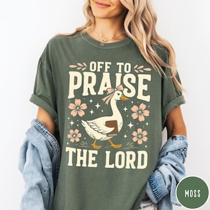 Puede incluir: Camiseta verde musgo con el texto "OFF TO PRAISE THE LORD" y un dibujo de un ganso con lazo y libro. El dise&ntilde;o incluye flores rosas y estrellas. La camiseta es de corte holgado.