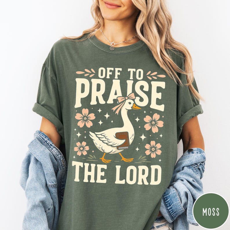 Puede incluir: Camiseta verde musgo con el texto "OFF TO PRAISE THE LORD" y un dibujo de un ganso con lazo y libro. El dise&ntilde;o incluye flores rosas y estrellas. La camiseta es de corte holgado.