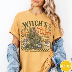 May include: A mustard yellow t-shirt featuring a graphic print with the text Witch's Herb Garden, Freshly Gathered, Locally Grown Herbs, Moonlit Grown, Autumn Picked, Sage, Potions, Moonlight, and a bottle labeled Witch's Brew with herbs and mushrooms.