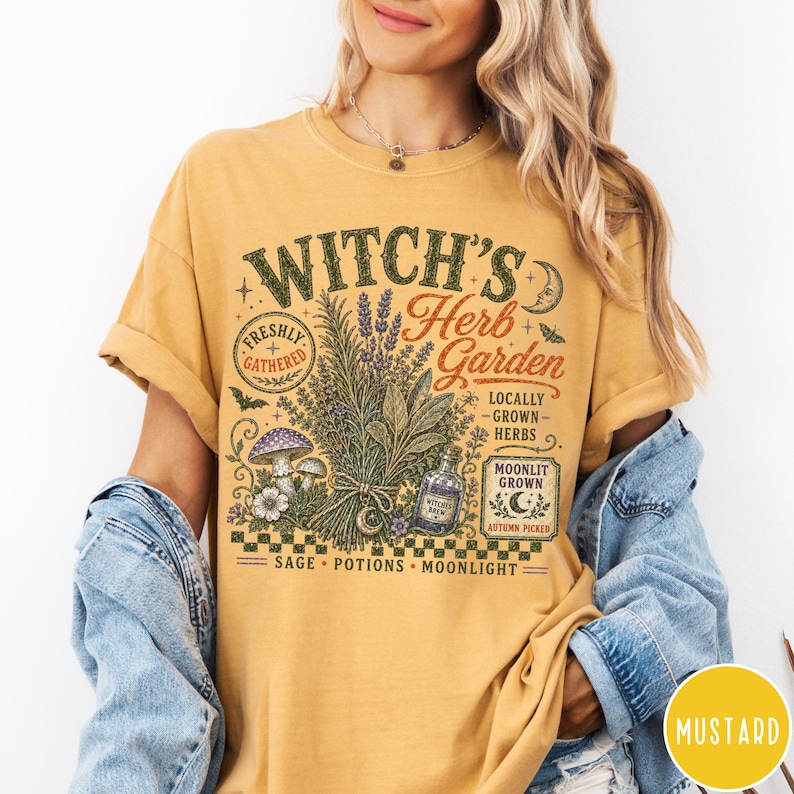 May include: A mustard yellow t-shirt featuring a graphic print with the text Witch's Herb Garden, Freshly Gathered, Locally Grown Herbs, Moonlit Grown, Autumn Picked, Sage, Potions, Moonlight, and a bottle labeled Witch's Brew with herbs and mushrooms.