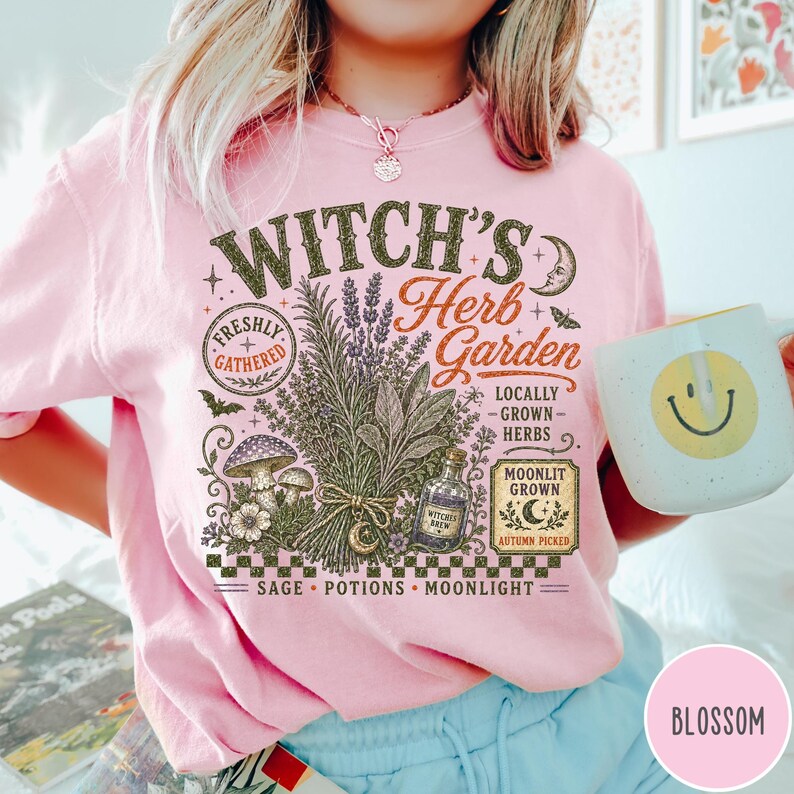 May include: A light pink t-shirt featuring a graphic print with the text Witch's Herb Garden. The design includes illustrations of a bundle of herbs, mushrooms, a potion bottle, a crescent moon, and stars, with the words Sage, Potions, Moonlight.