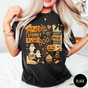 Op de afbeelding: Zwart T-shirt met een Halloween-thema. Het ontwerp bevat pompoenen, geesten, een heksenhoed en de zinnen "Spooky Boo!" en "Happy Halloween". Het shirt heeft een ronde hals en korte mouwen.