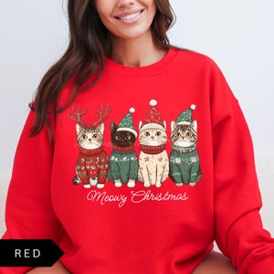 May include: Red sweatshirt featuring four cats in Christmas sweaters and hats. The cats are wearing green and red sweaters and hats, with one cat wearing antlers. The text "Meowy Christmas" is written below the cats.