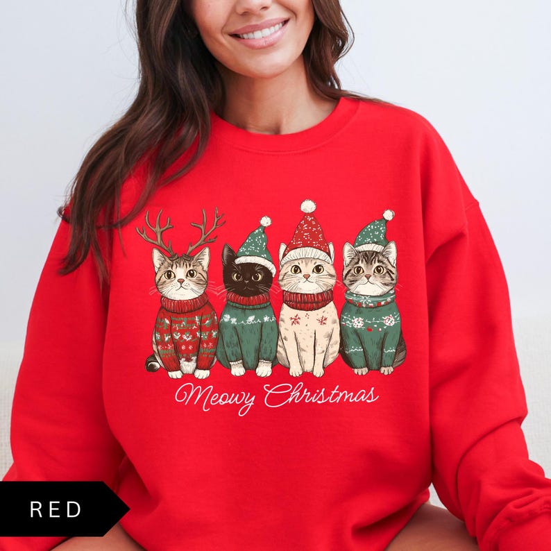 May include: Red sweatshirt featuring four cats in Christmas sweaters and hats. The cats are wearing green and red sweaters and hats, with one cat wearing antlers. The text "Meowy Christmas" is written below the cats.