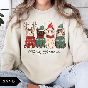 May include: Sand-colored sweatshirt featuring four cats in festive sweaters and hats. The cats are adorned with reindeer antlers and Santa hats. The text "Meowy Christmas" is written in a script font below the cats. The word "SAND" is in a black box at the bottom left.