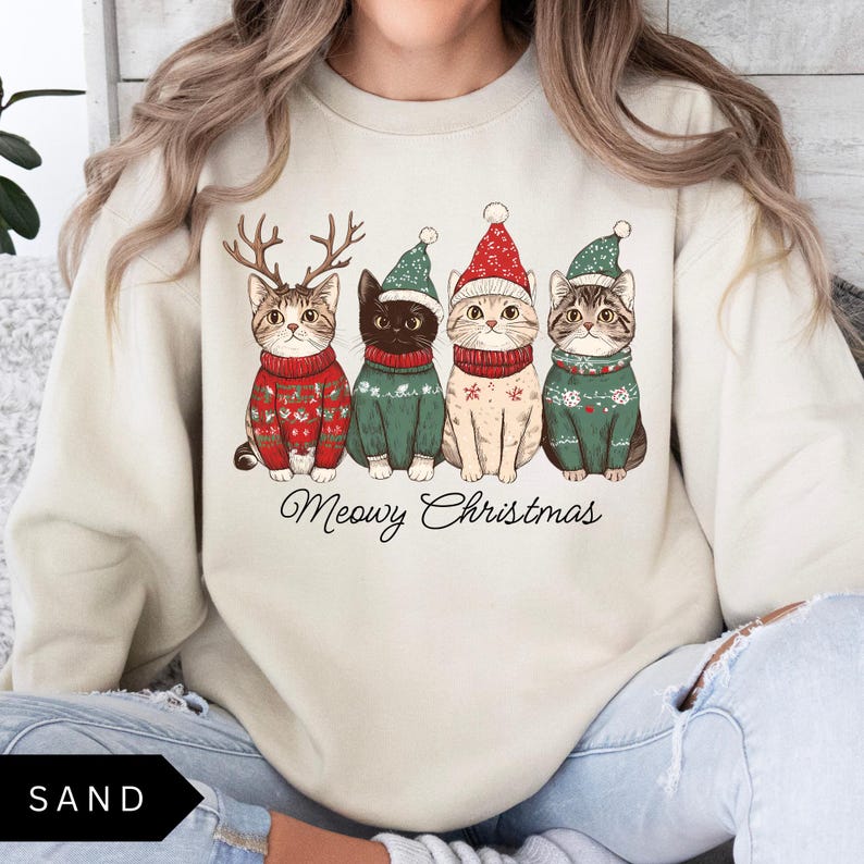 May include: Sand-colored sweatshirt featuring four cats in festive sweaters and hats. The cats are adorned with reindeer antlers and Santa hats. The text "Meowy Christmas" is written in a script font below the cats. The word "SAND" is in a black box at the bottom left.