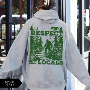 May include: A gray hooded sweatshirt with the text "Respect the Locals" printed on the back. The design features a green forest scene with a Bigfoot figure walking through the trees.