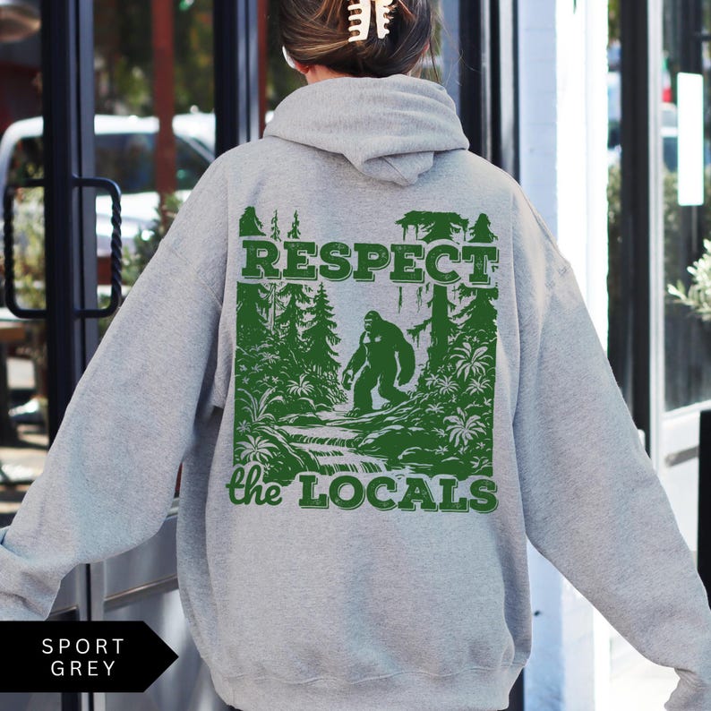 May include: A gray hooded sweatshirt with the text "Respect the Locals" printed on the back. The design features a green forest scene with a Bigfoot figure walking through the trees.