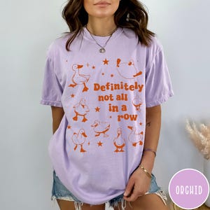 May include: A lavender t-shirt with a graphic of ducks and stars. The text "Definitely not all in a row" is printed in orange on the shirt.