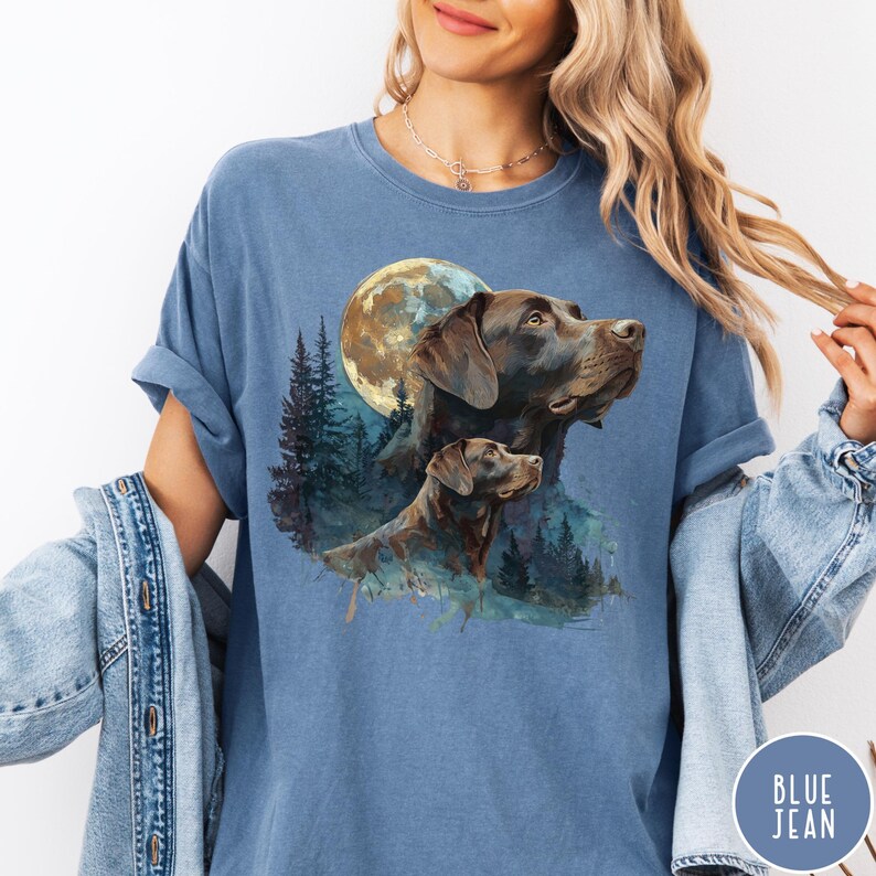 May include: Blue t-shirt featuring a watercolor illustration of two brown dogs with a full moon and forest background. The shirt has rolled-up sleeves and a relaxed fit. A blue circle with the words "BLUE JEAN" is in the bottom right corner.