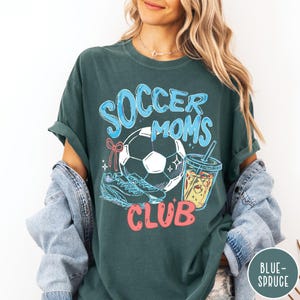 May include: Dark teal t-shirt with the words "Soccer Moms Club" in blue and red. The graphic includes a soccer ball, soccer cleats, and a drink with a straw. The shirt is a comfortable, oversized fit.