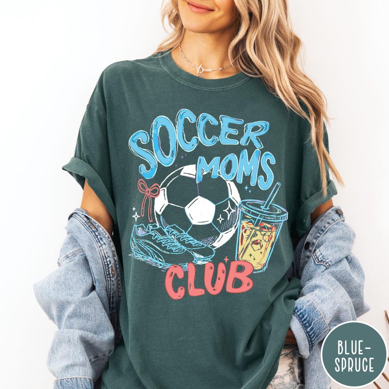 May include: Dark teal t-shirt with the words "Soccer Moms Club" in blue and red. The graphic includes a soccer ball, soccer cleats, and a drink with a straw. The shirt is a comfortable, oversized fit.