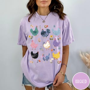 K&ouml;nnte beinhalten: Ein lavendelfarbenes T-Shirt mit einer bunten Grafik von H&uuml;hnern. Die H&uuml;hner sind in verschiedenen Posen und haben unterschiedlich farbige Federn. Das Shirt hat eine lockere Passform und einen Rundhalsausschnitt.